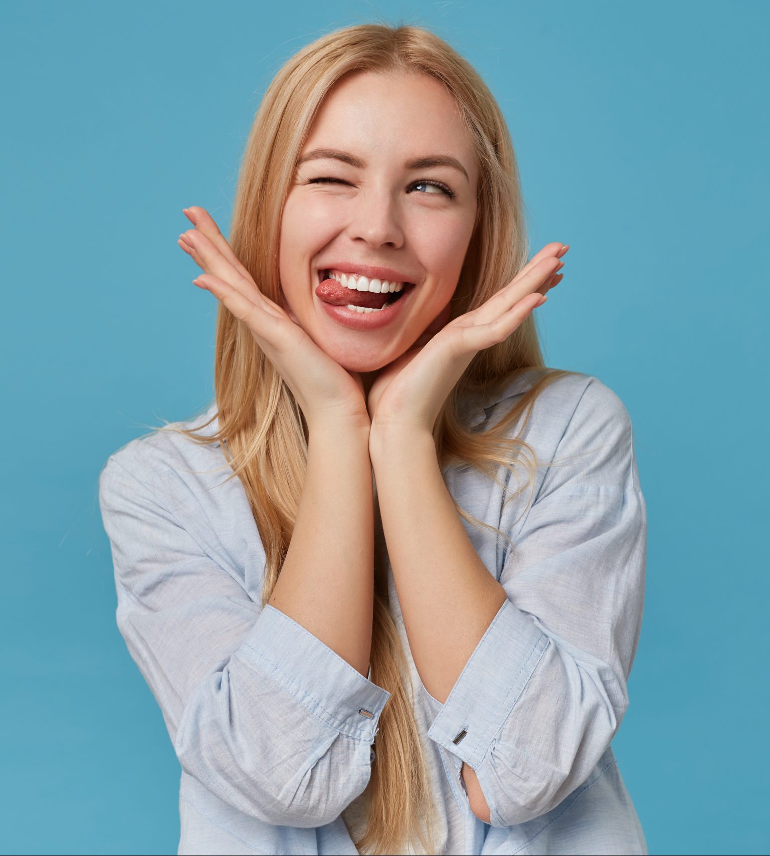 Portrait of pretty young blonde lady with long hair keeping head on raised hands, making faces over blue background, looking aside joyfully with closed eye and showinng tongue