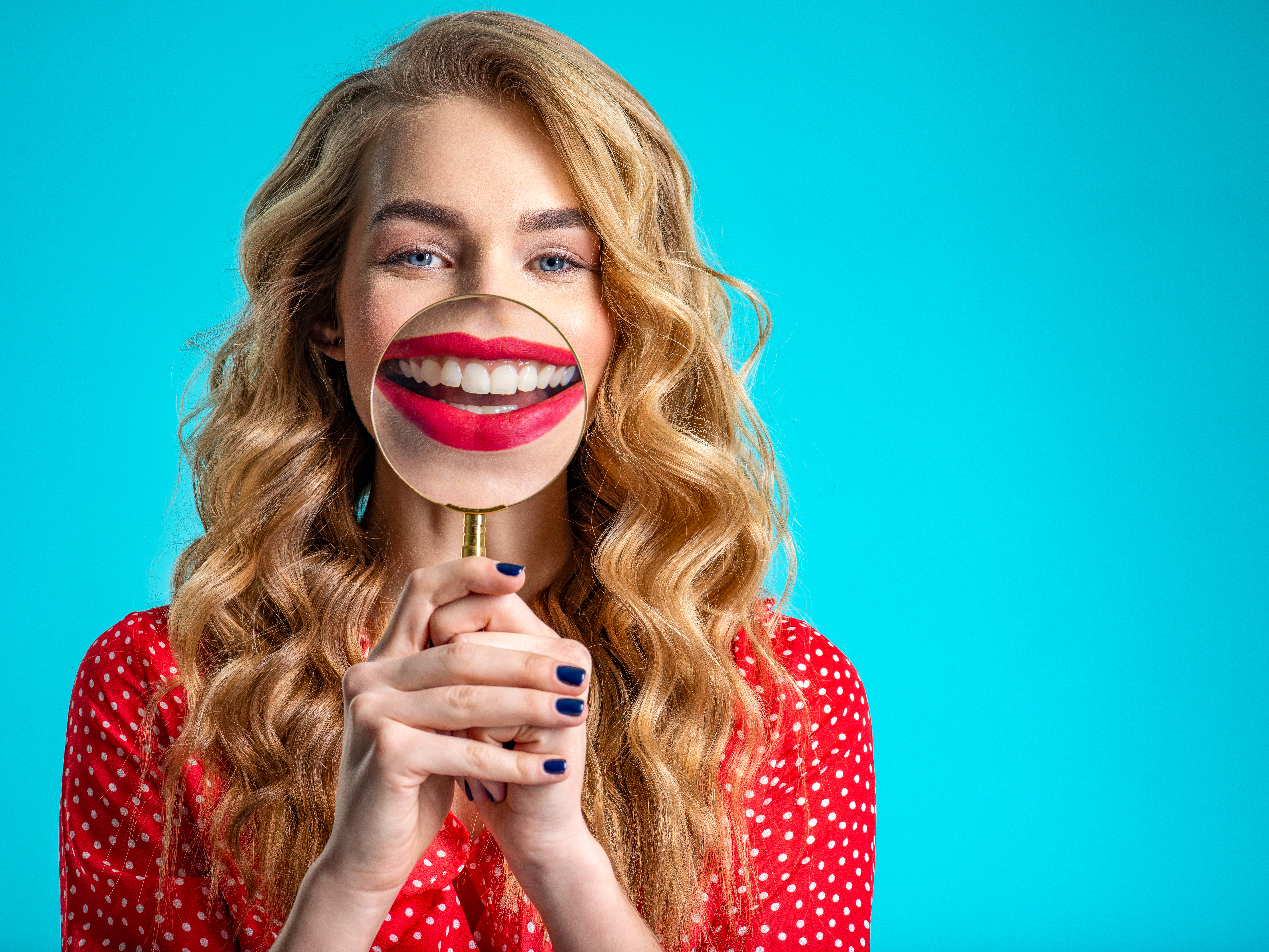 Blonde holds a magnifying glass in front of her teeth. Girl with