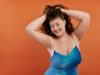 A beautiful young woman in a blue dress playfully shows her confidence on a vibrant orange backdrop.