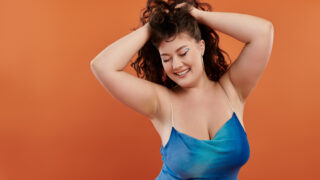 A beautiful young woman in a blue dress playfully shows her confidence on a vibrant orange backdrop.